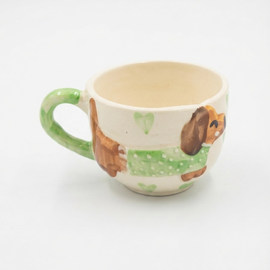 A small ceramic teacup with a green handle and a hand-painted dachshund design wearing a green sweater, set against a plain white background.