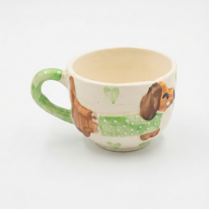 A small ceramic teacup with a green handle and a hand-painted dachshund design wearing a green sweater, set against a plain white background.