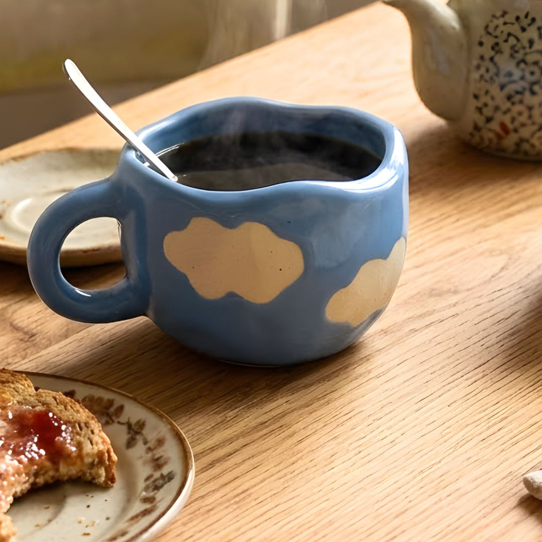 Blue cloud-shaped mug with steaming liquid on a wooden table with a teapot and plate in the background.