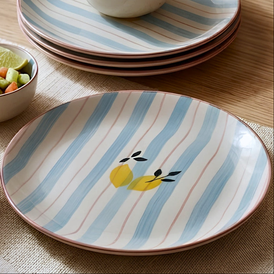 A set of ceramic dinnerware on a wooden table, featuring round plates and bowls with light blue and white vertical stripes, a pink rim, and a yellow lemon illustration on the front plate.