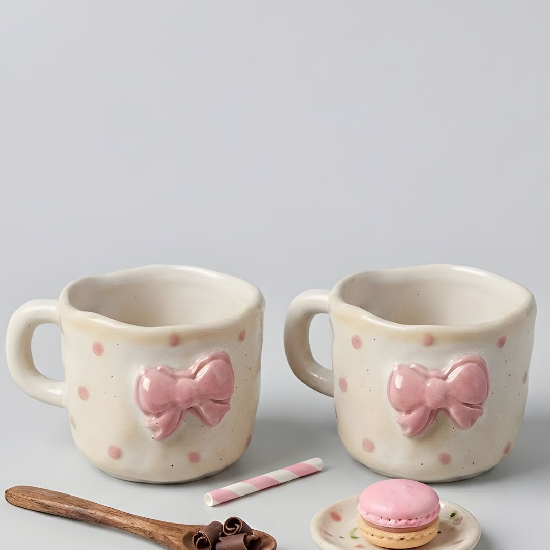 Two ceramic mugs with pink bow designs on a gray background, accompanied by a wooden spoon and small dish with a macaron.
