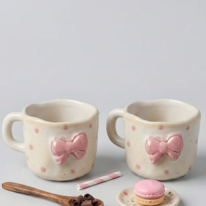 Two ceramic mugs with pink bow designs on a gray background, accompanied by a wooden spoon and small dish with a macaron.