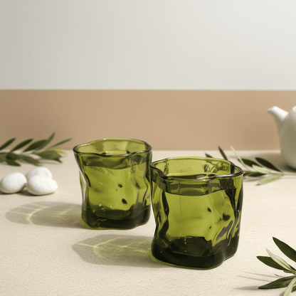 Two green glass tumblers on a beige surface with white stones and green leaves.