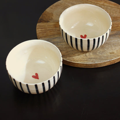 Two ceramic bowls with black stripes and red hearts on a wooden board against a black background.