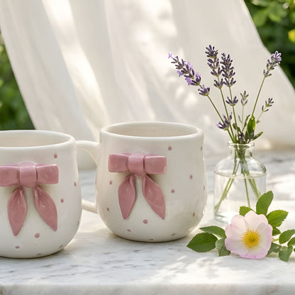 Two ceramic mugs with pink bows on a wooden surface with flowers and fabric in the background.