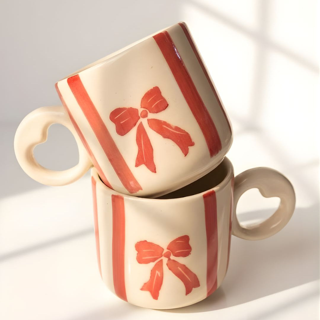 Two ceramic mugs with red bow designs stacked on a light background