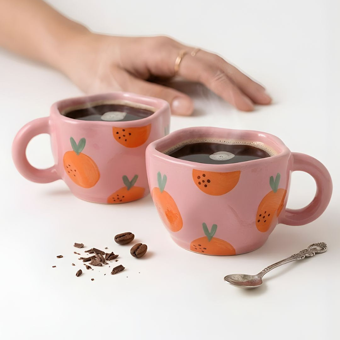 Two pink mugs with orange and green designs on a white surface, filled with a dark liquid.