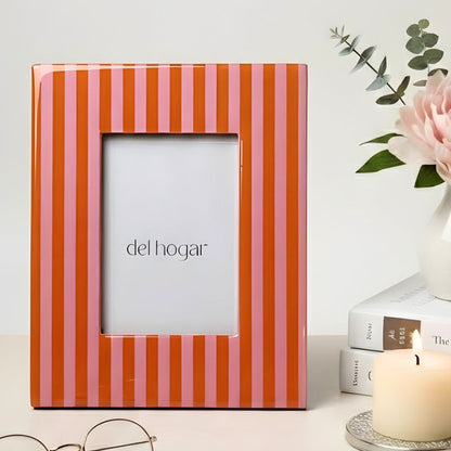 Striped photo frame with 'del hogar' branding, next to a vase with a pink flower, books, and glasses on a light surface.