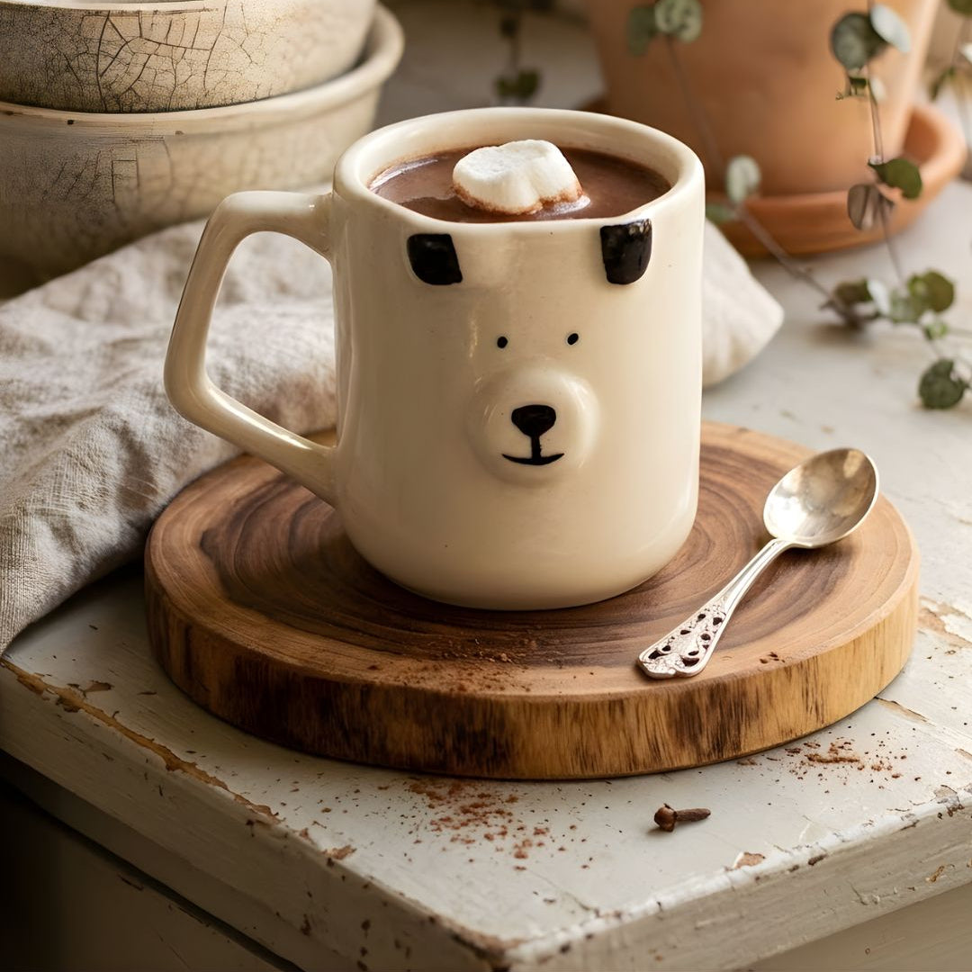 White ceramic mug with bear face design on a wooden coaster, filled with hot chocolate and marshmallows.