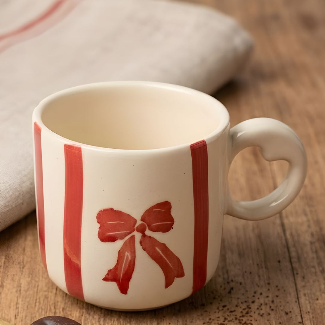 White mug with red bow design on a wooden surface with coffee beans and a spoon.