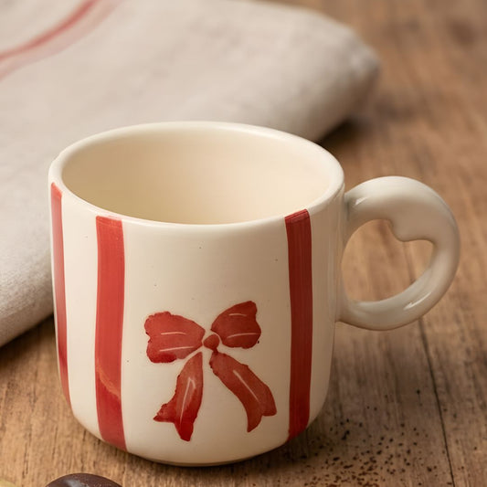 White mug with red bow design on a wooden surface with coffee beans and a spoon.
