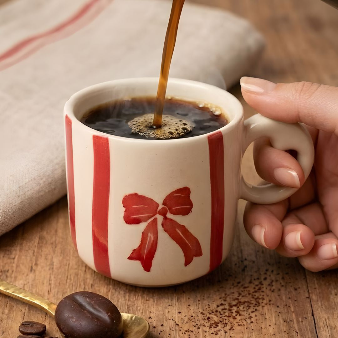 Person pouring coffee into a decorative mug with a red bow design on a wooden surface.