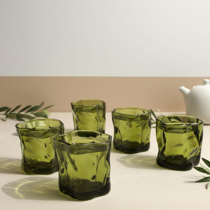 Set of six green glass tumblers on a light surface with a white teapot in the background.