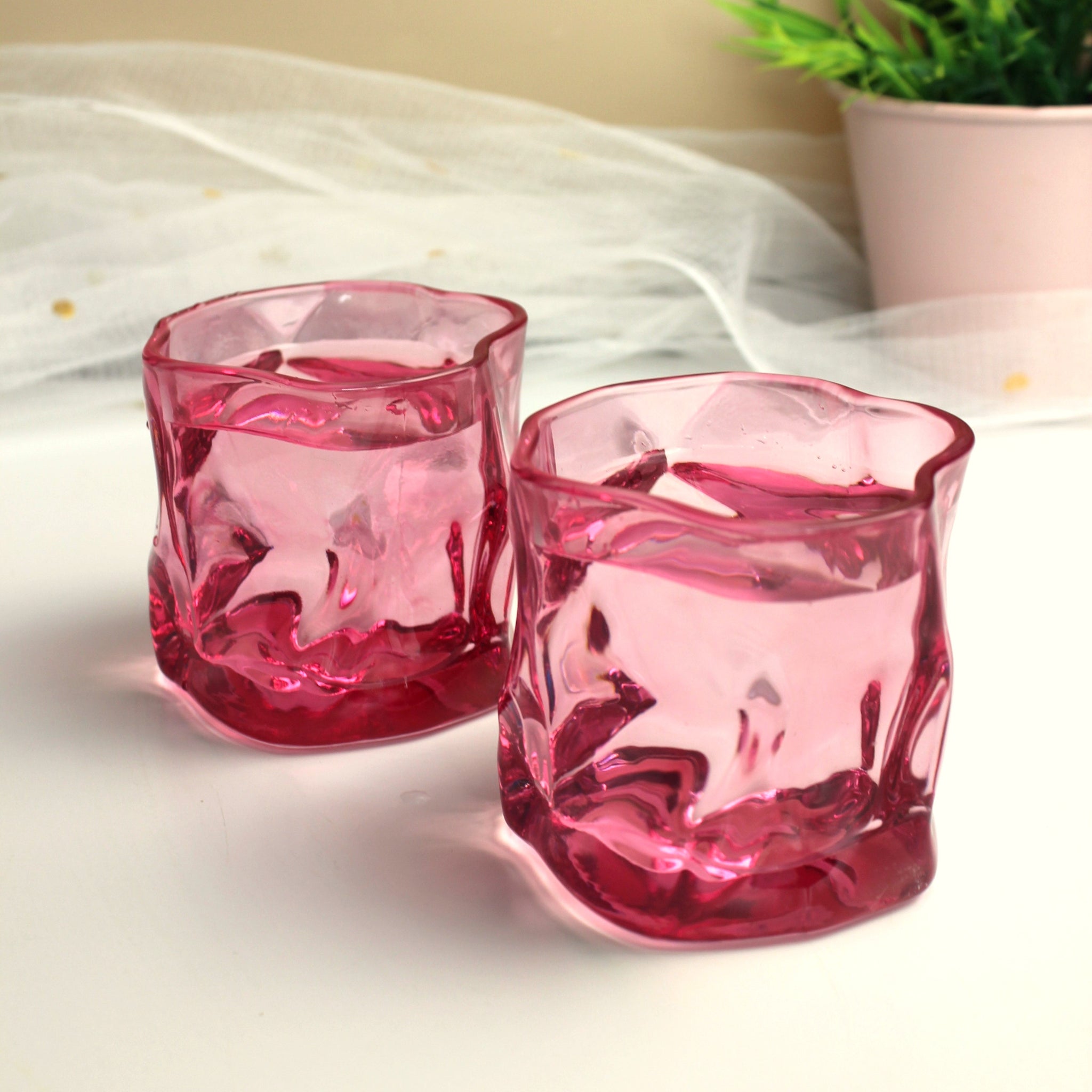 Two pink glass votive holders on a white surface with a blurred plant in the background.