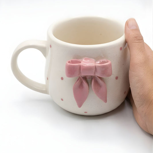 Ceramic mug with a pink bow design held by a hand on a white background